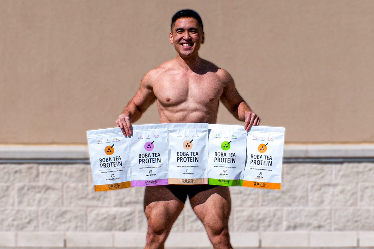 Shirtless man smiling and holding four large bags of Boba Tea Protein in front of a beige wall.