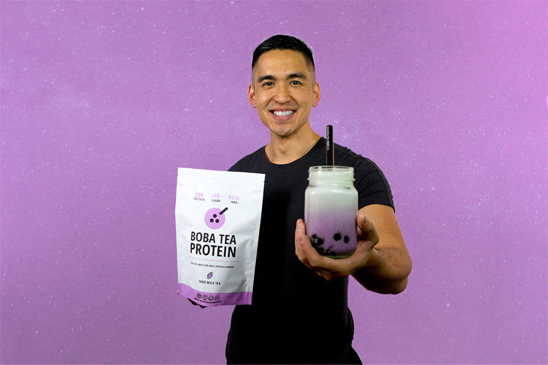 Man smiling while holding a Taro Boba Tea Protein bag and a matching boba drink in front of a purple surface.