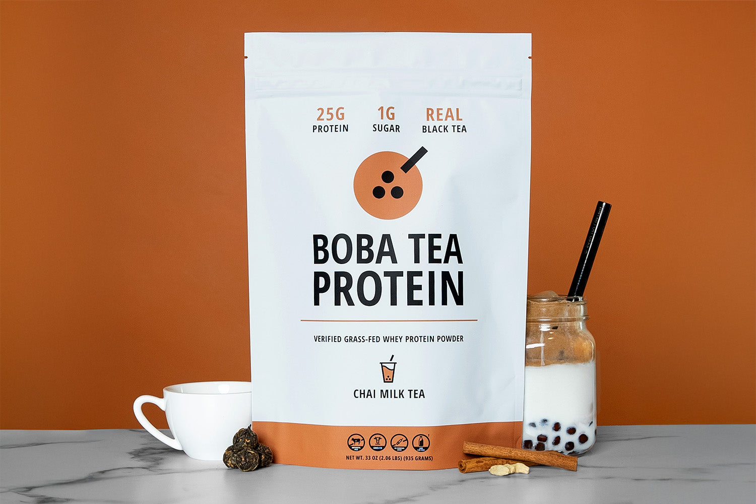 The image shows a bag of "Boba Tea Protein" powder in Chai Milk Tea flavor, featuring highlights like "25g Protein", "1g Sugar", and "Real Black Tea", against a background of light brown matching the chai theme. In front are tea leaves, cinnamon sticks and cardamom seeds, a teacup, and a jar of Boba Tea Protein's Chai Milk Tea poured into milk with tapioca pearls and a metal straw.