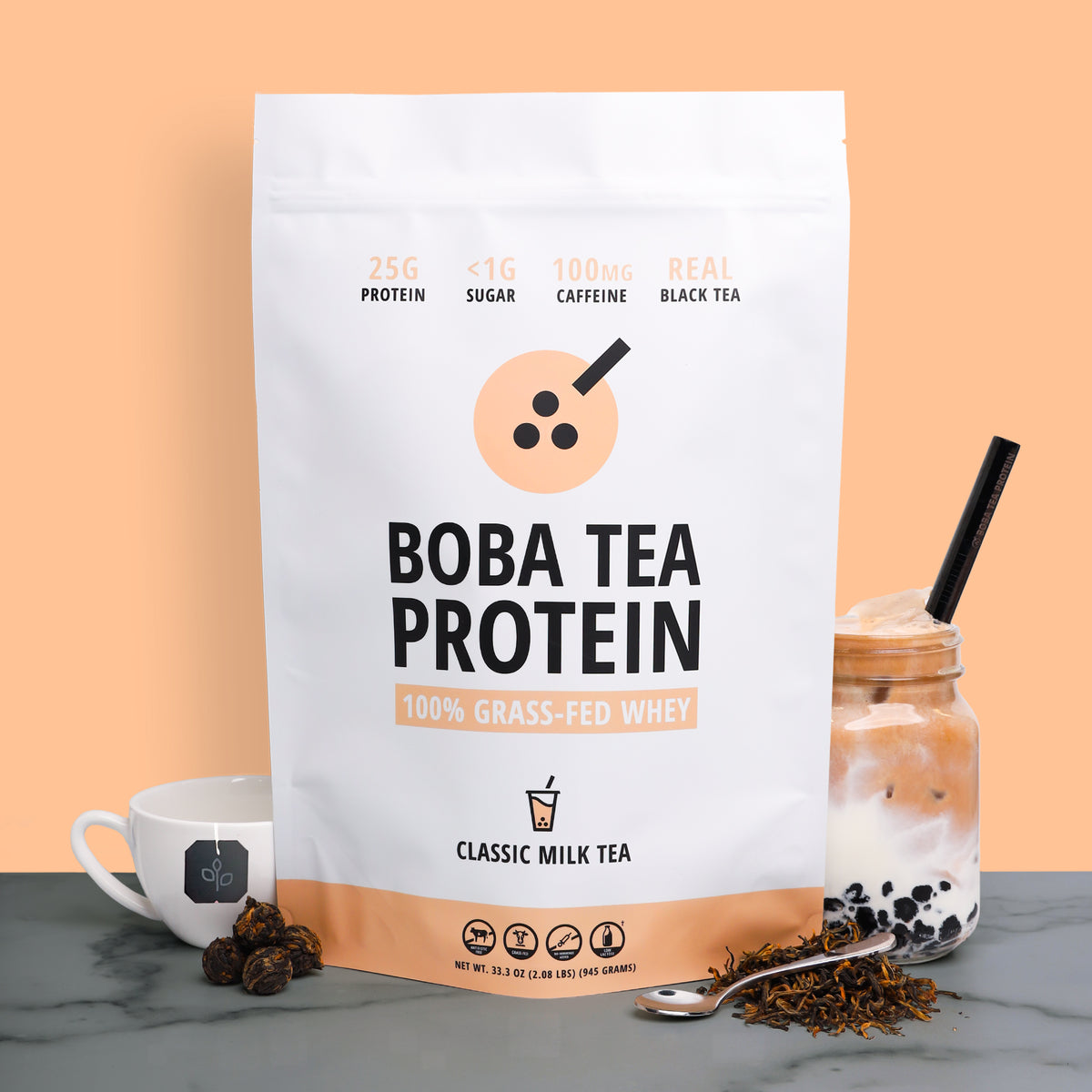 A stand-up pouch of Boba Tea Protein powder stands on a gray marble surface against a peach background, styled with a white tea cup, a boba milk tea drink in a glass jar, loose tea leaves, and a spoon.