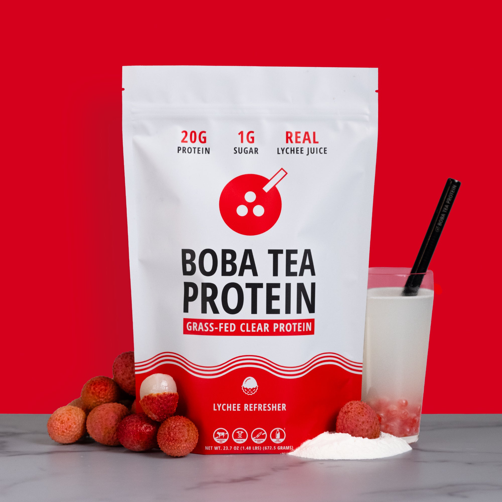 A stand-up pouch of Boba Tea Protein grass-fed clear protein in Lychee Refresher flavor, shown on a gray surface with lychee fruits, a glass of white drink with red popping pearls, and a black straw against a solid red background.