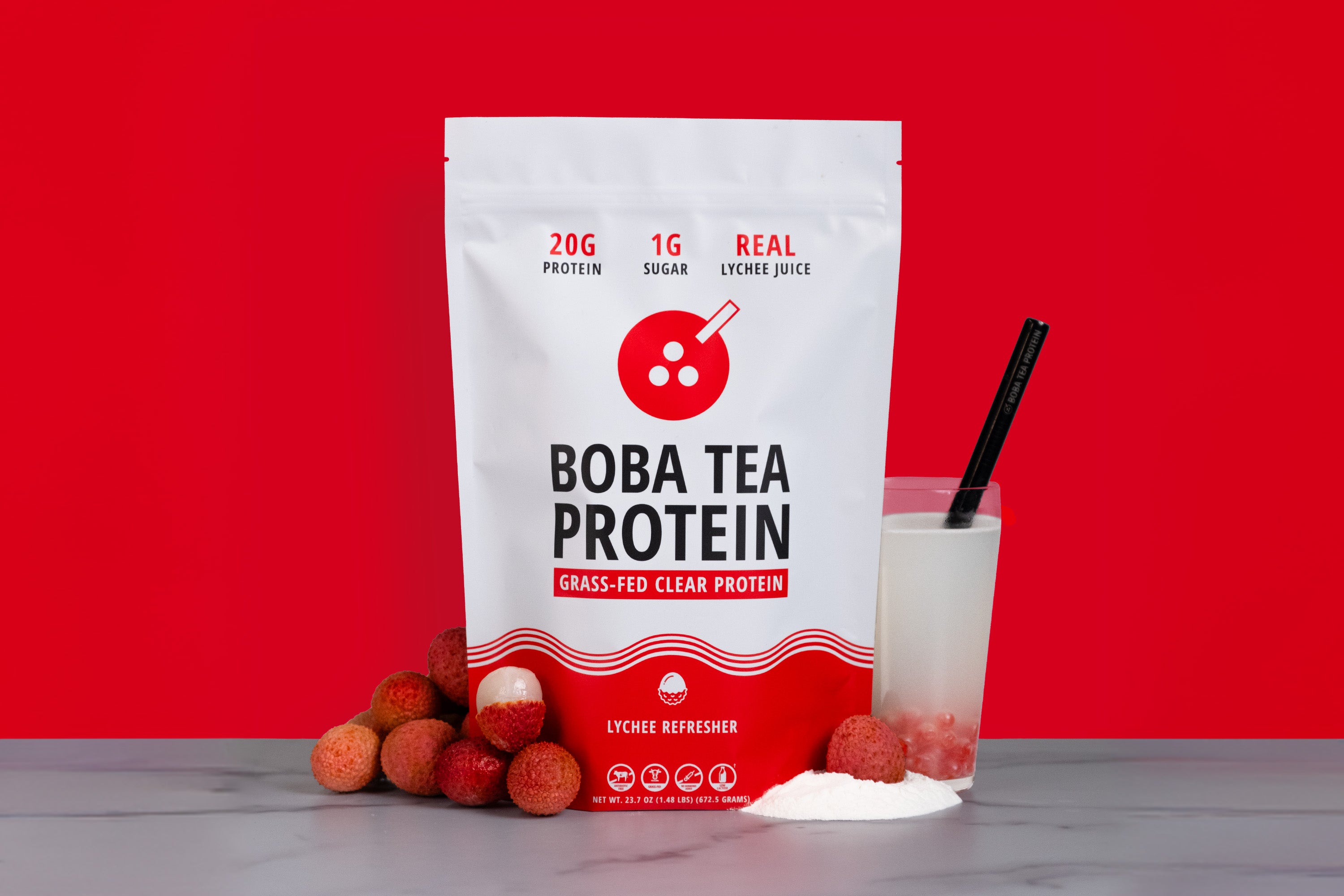 A stand-up pouch of Boba Tea Protein grass-fed clear protein powder, shown with a glass of white drink and lychees on a marble surface against a bright red background.