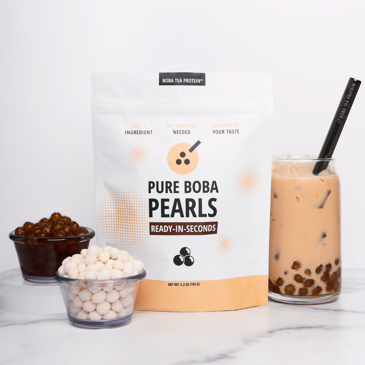 A stand-up pouch of Pure Boba Pearls stands on a white marble surface, with two small bowls of boba pearls, one dry and one soaked in syrup, and a glass of iced milk tea with a black straw. The pouch is white with peach and black text and graphics.