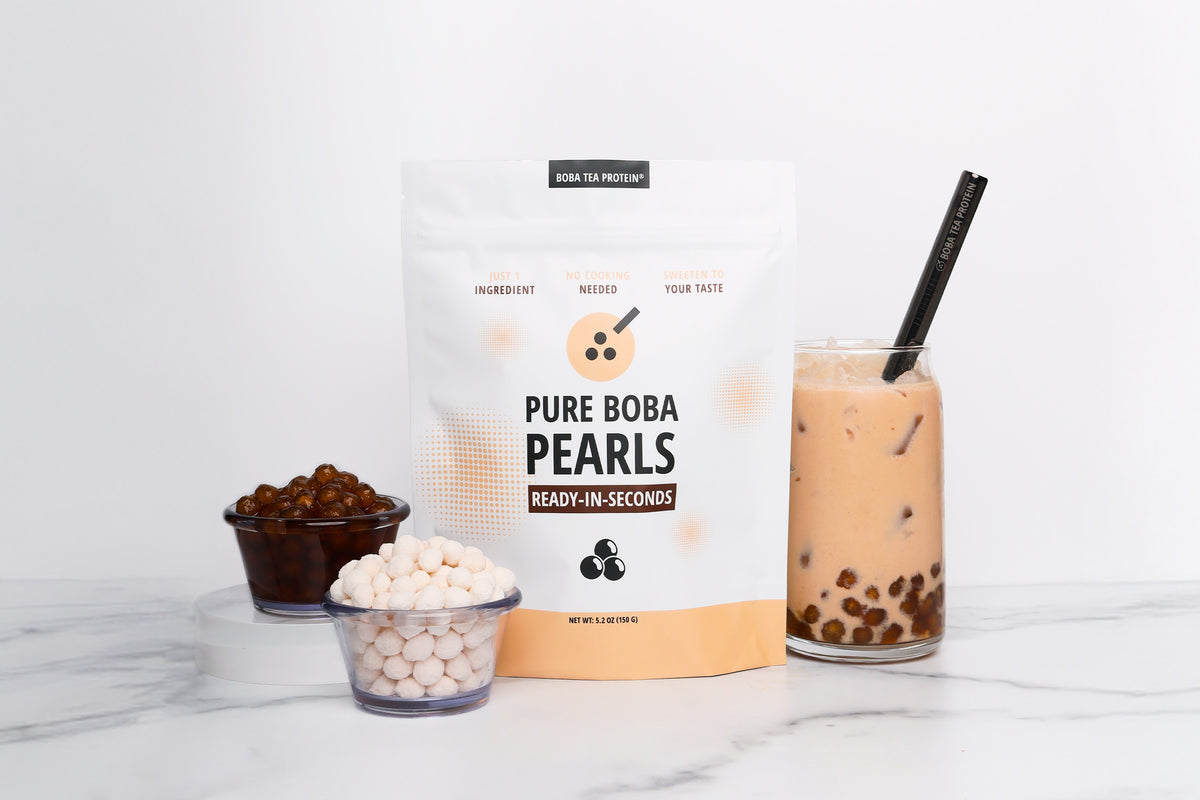 A bag of Boba Tea Protein Pure Boba Pearls stands on a white marble surface next to two small bowls containing the pearls, one dry and one soaked in syrup, and a glass of a milk tea drink with boba pearls and a black straw.