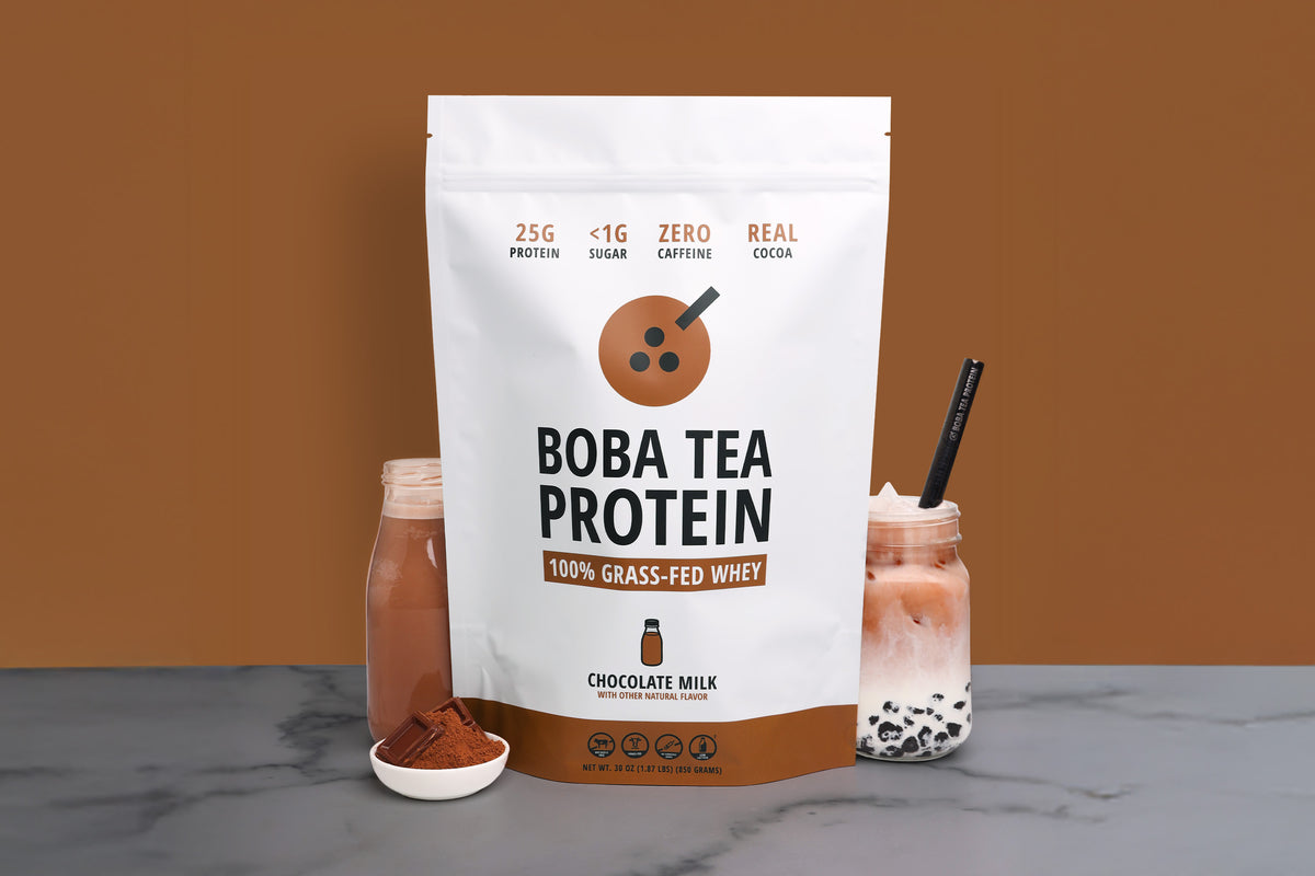 A 25-serving stand-up pouch of Boba Tea Protein Chocolate Milk whey powder stands on a gray marble surface against a solid brown background. The white and brown packaging features a milk bottle graphic and text indicating 25g protein, <1g sugar, zero caffeine, and real cocoa.