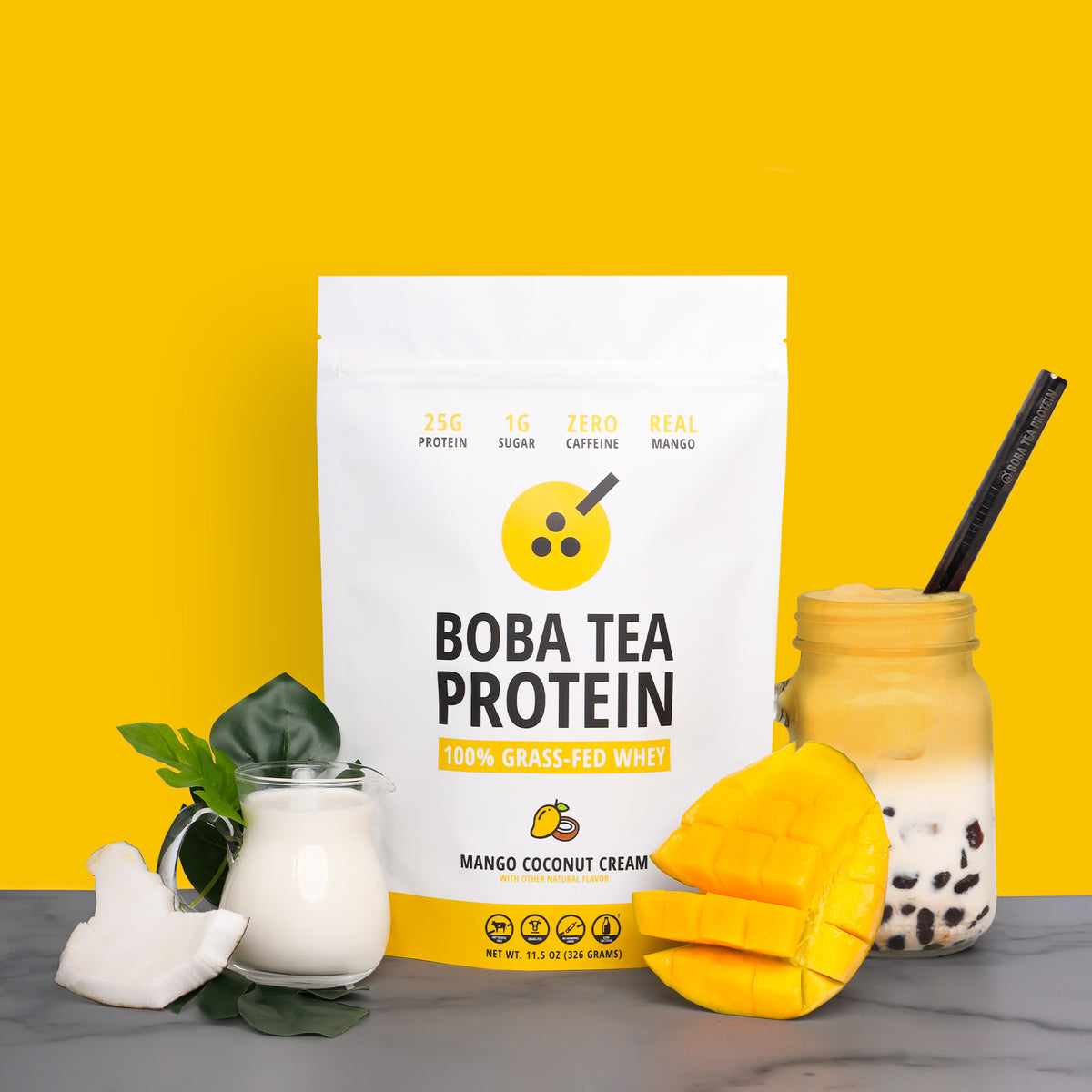 A bag of Boba Tea Protein powder stands on a marble surface against a bright yellow background, styled with a glass jar of boba tea, mango slices, coconut, and a small glass of coconut milk.