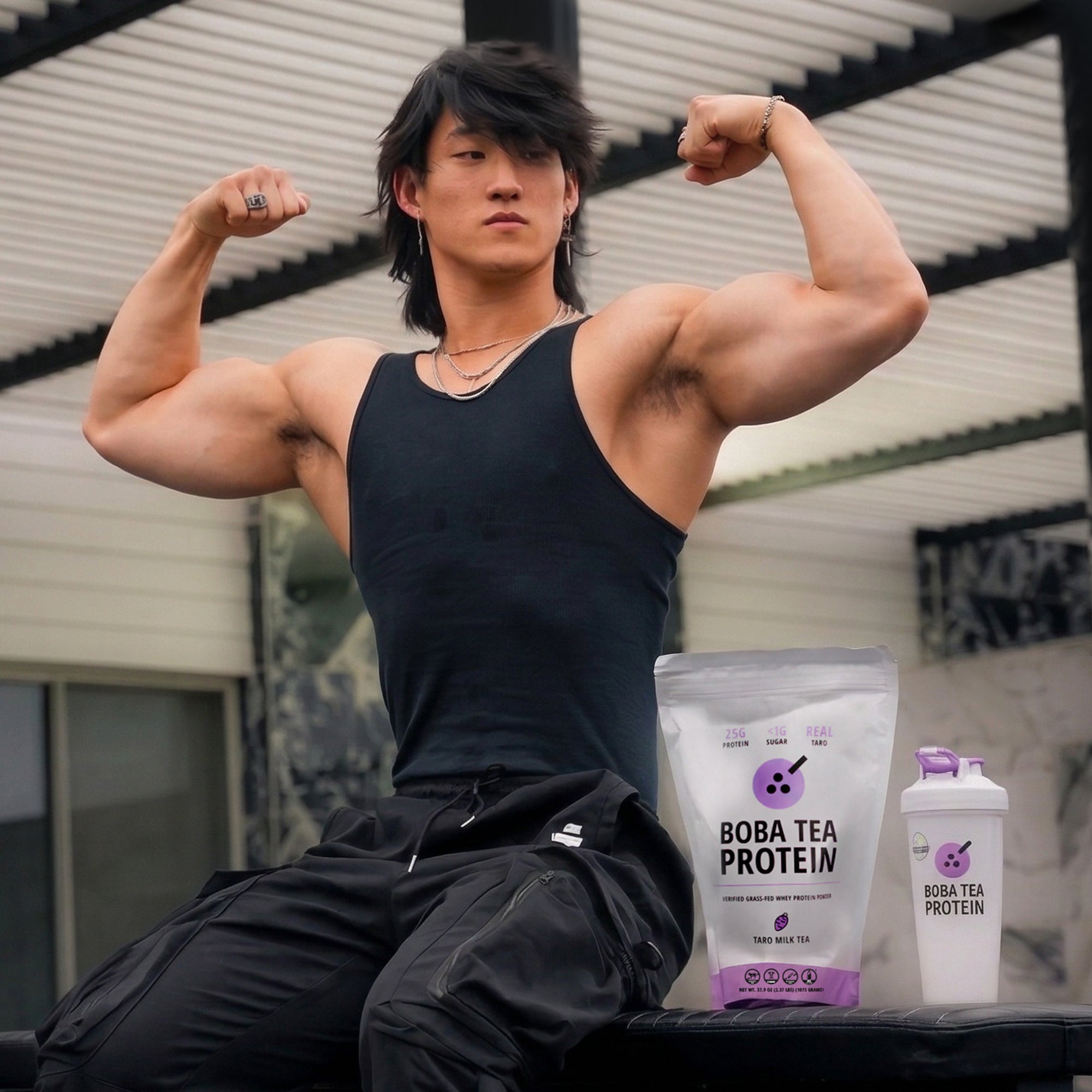 A muscular man in a black tank top poses flexing his arms outdoors, next to a bag of Taro Milk Tea Whey Protein from Boba Tea Protein and a Boba Tea Protein branded shaker bottle in matching Taro purple.