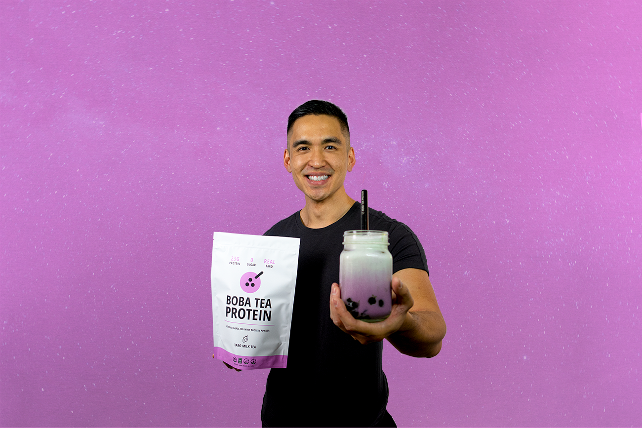 Man smiling while holding a Taro Boba Tea Protein bag and a matching boba drink in front of a purple surface.