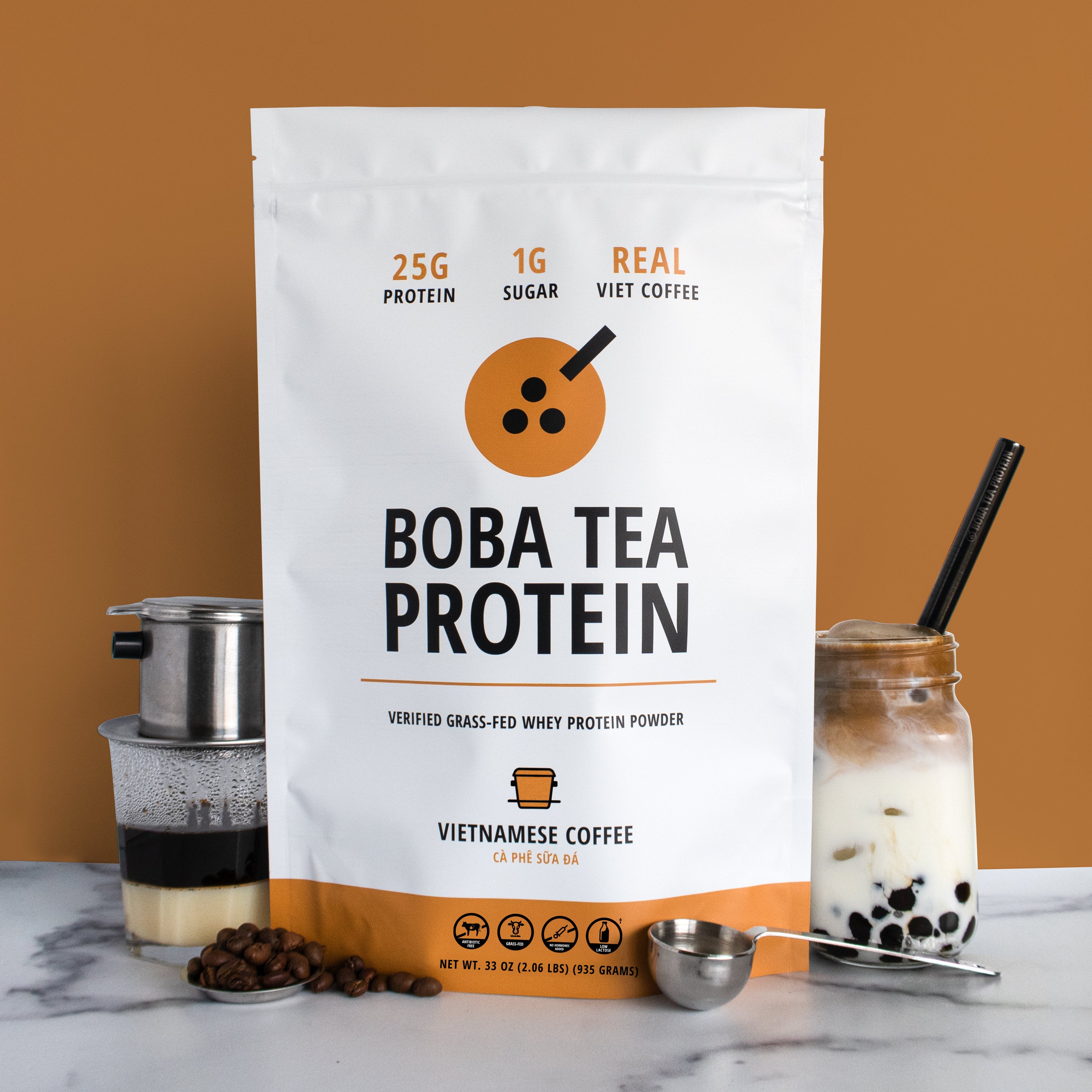 The image shows a bag of "Boba Tea Protein" powder in Vietnamese Coffee flavor, featuring highlights like "25g Protein" and "1g Sugar", against a background of light brown matching the coffee theme. In front are coffee beans, a French drip press over a cup of brewed coffee and condensed milk, a metal scoop, and a jar of Boba Tea Protein's Vietnamese Coffee poured into milk with tapioca pearls and a metal straw.