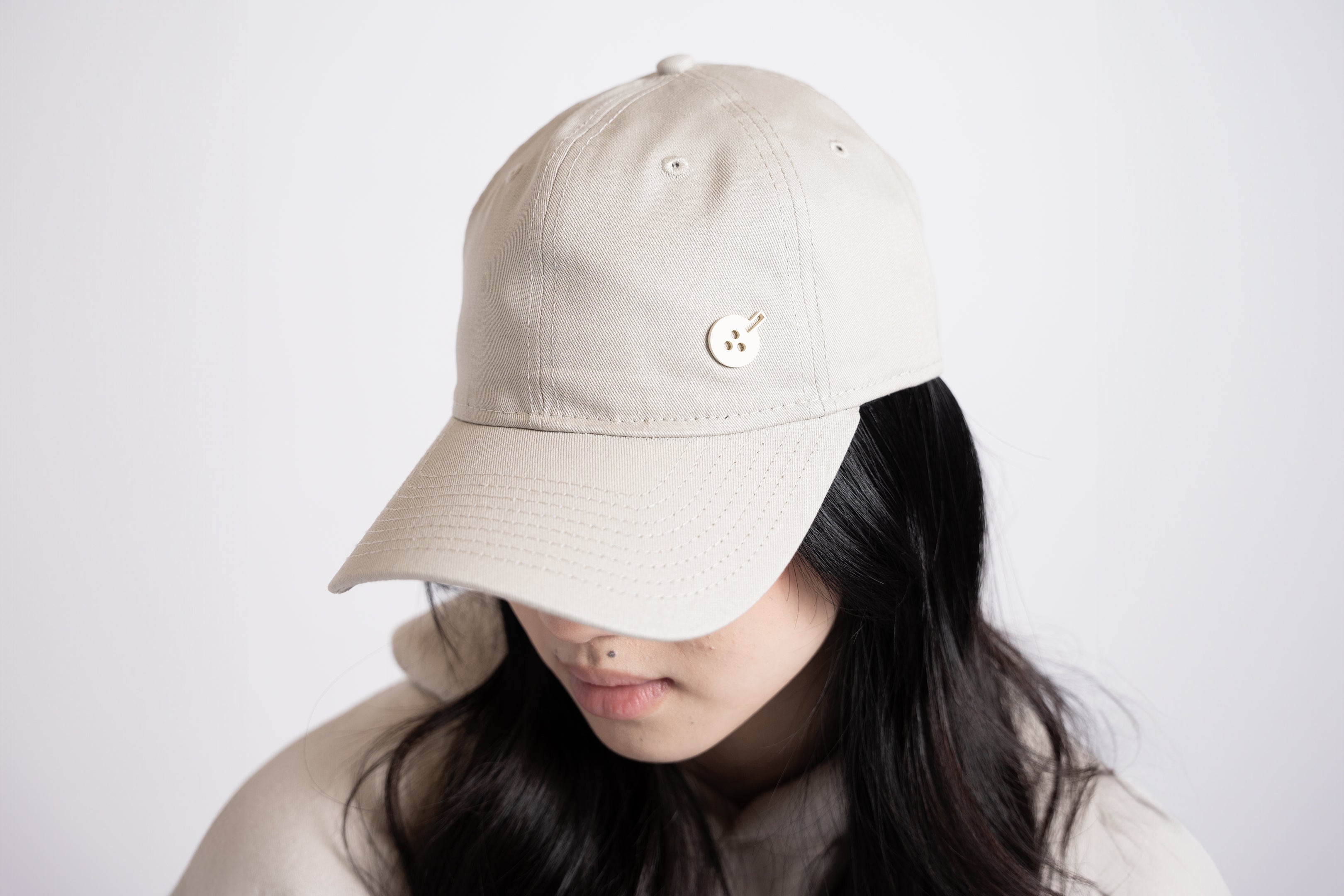 A beige cloth hat in "dad hat" style, with a silicone embellishment of the same color in the shape of Boba Tea Protein's circular logo depicting a boba drink placed to the side of the front of the hat.