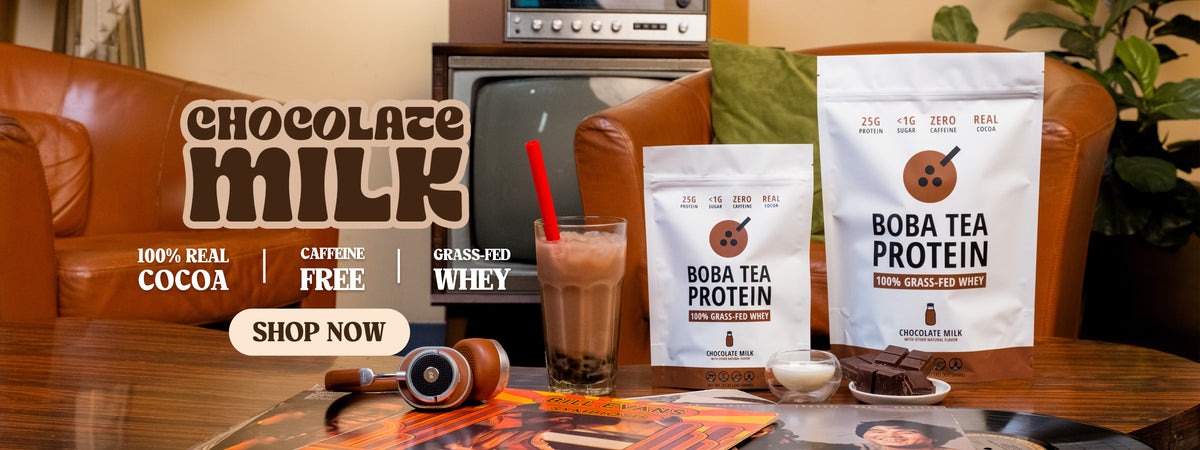 Two bags of Boba Tea Protein powder are displayed on a wooden table in a cozy living room setting, with a glass of chocolate milk and a bowl of chocolate pieces in the foreground. The background features a brown leather sofa and a vintage television.