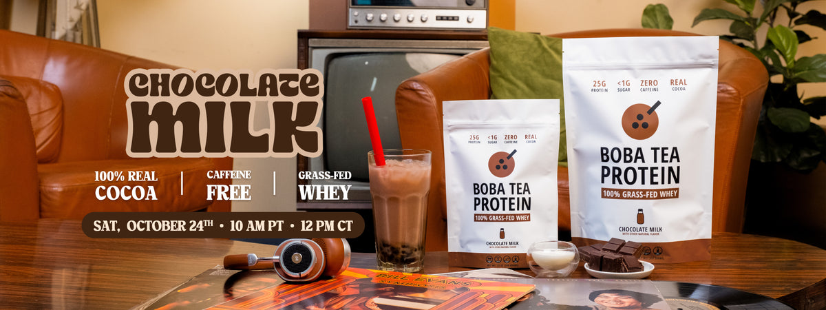Two bags of Boba Tea Protein Chocolate Milk powder are displayed on a wooden table in a cozy living room setting with a leather couch, a vintage television, and a glass of chocolate milk. The bags are white with brown, and the scene is overlaid with promotional text for the product.