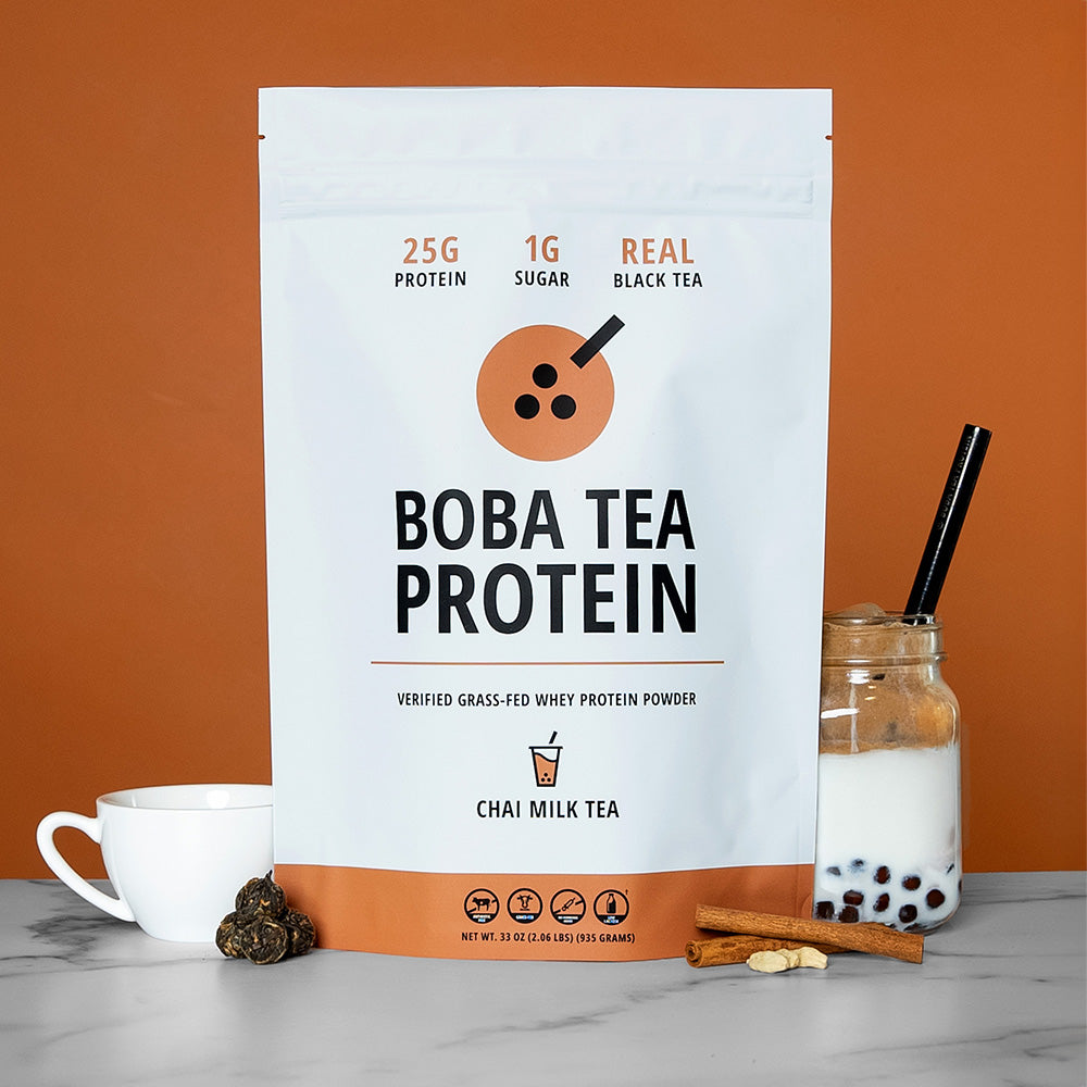 The image shows a bag of "Boba Tea Protein" powder in Chai Milk Tea flavor, featuring highlights like "25g Protein", "1g Sugar", and "Real Black Tea", against a background of light brown matching the chai theme. In front are tea leaves, cinnamon sticks and cardamom seeds, a teacup, and a jar of Boba Tea Protein's Chai Milk Tea poured into milk with tapioca pearls and a metal straw.