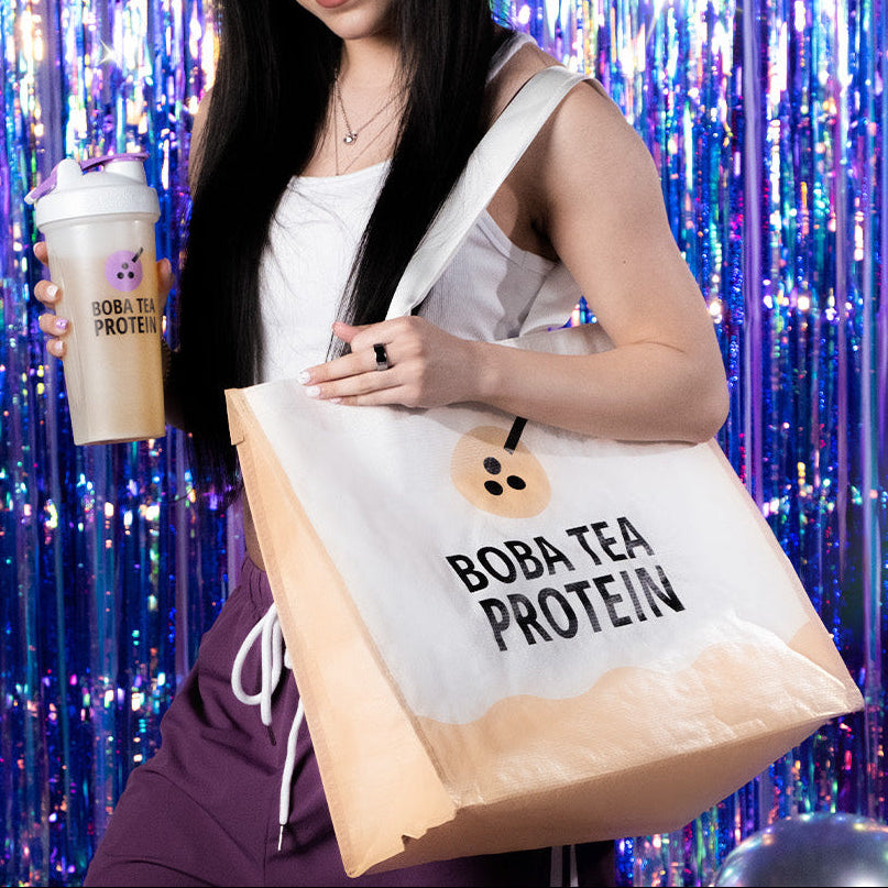Person holding a Boba Tea Protein cup and a matching tote bag against a colorful, tinsel-like background.