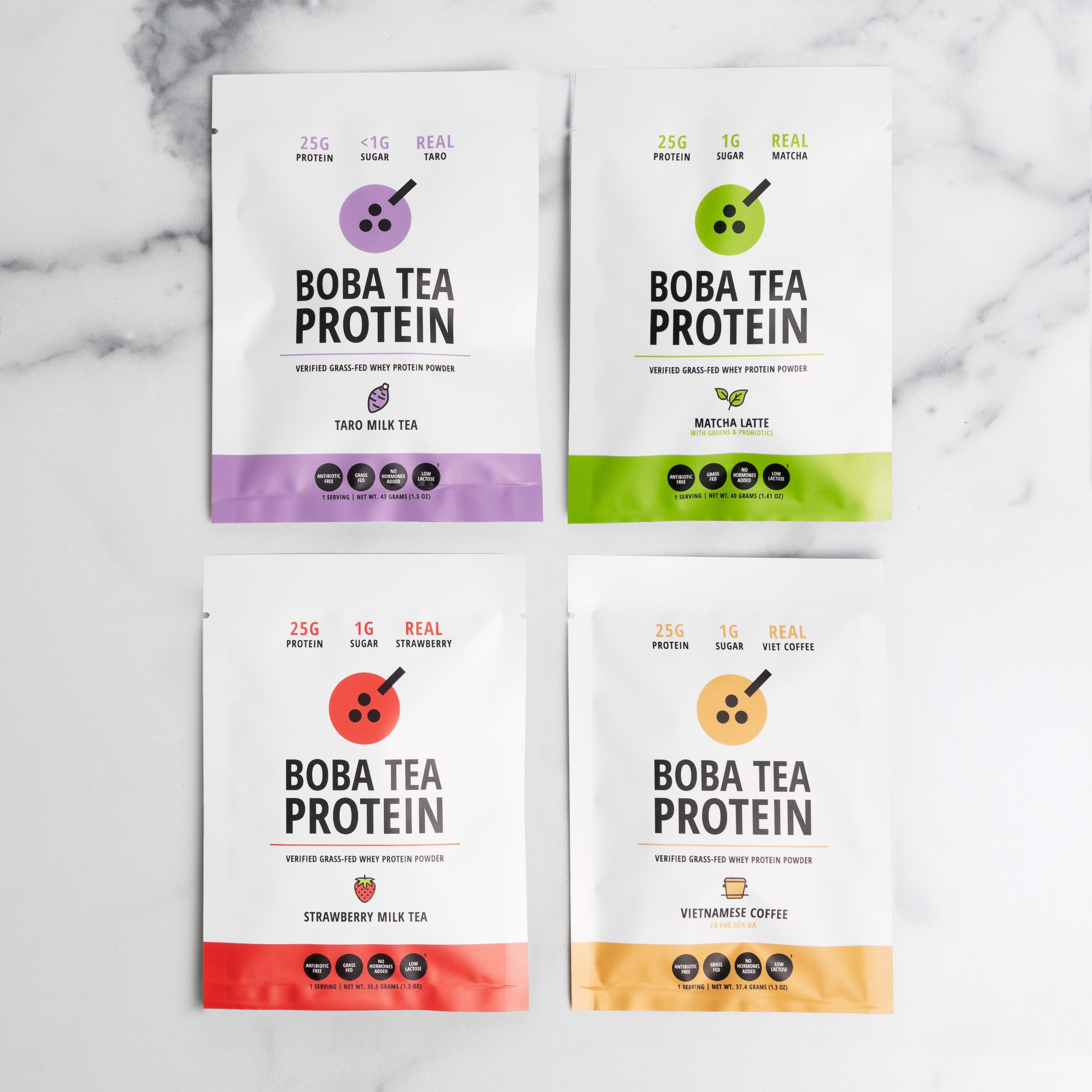 Four pouches of Boba Tea Protein powder arranged in a 2x2 grid on a light marble surface. The pouches are white with different colored accents and labels for Taro Milk Tea, Matcha Latte, Strawberry Milk Tea, and Vietnamese Coffee.