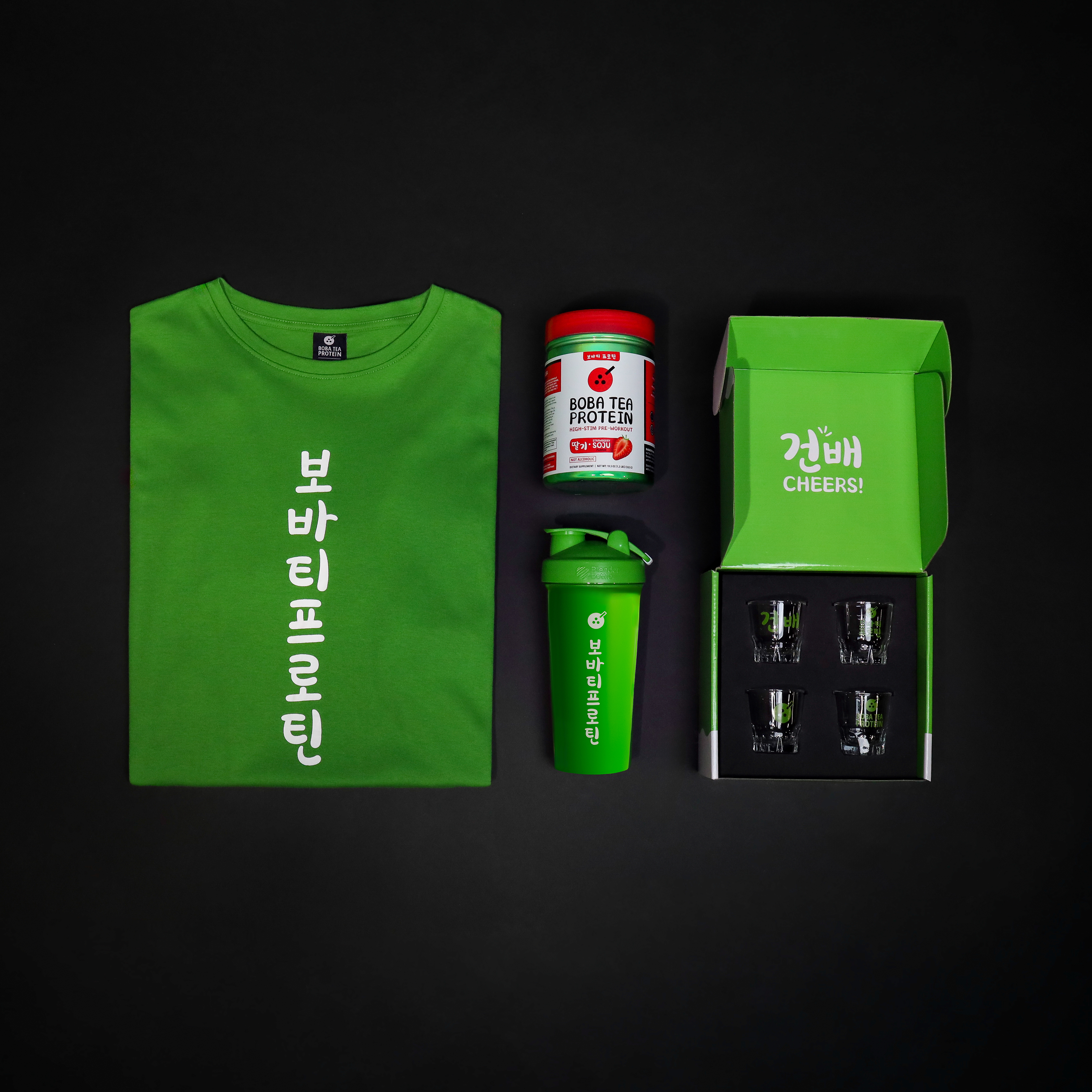 Green t-shirt, pre-workout supplement, shaker bottle, and box of glass shot cups on a black background