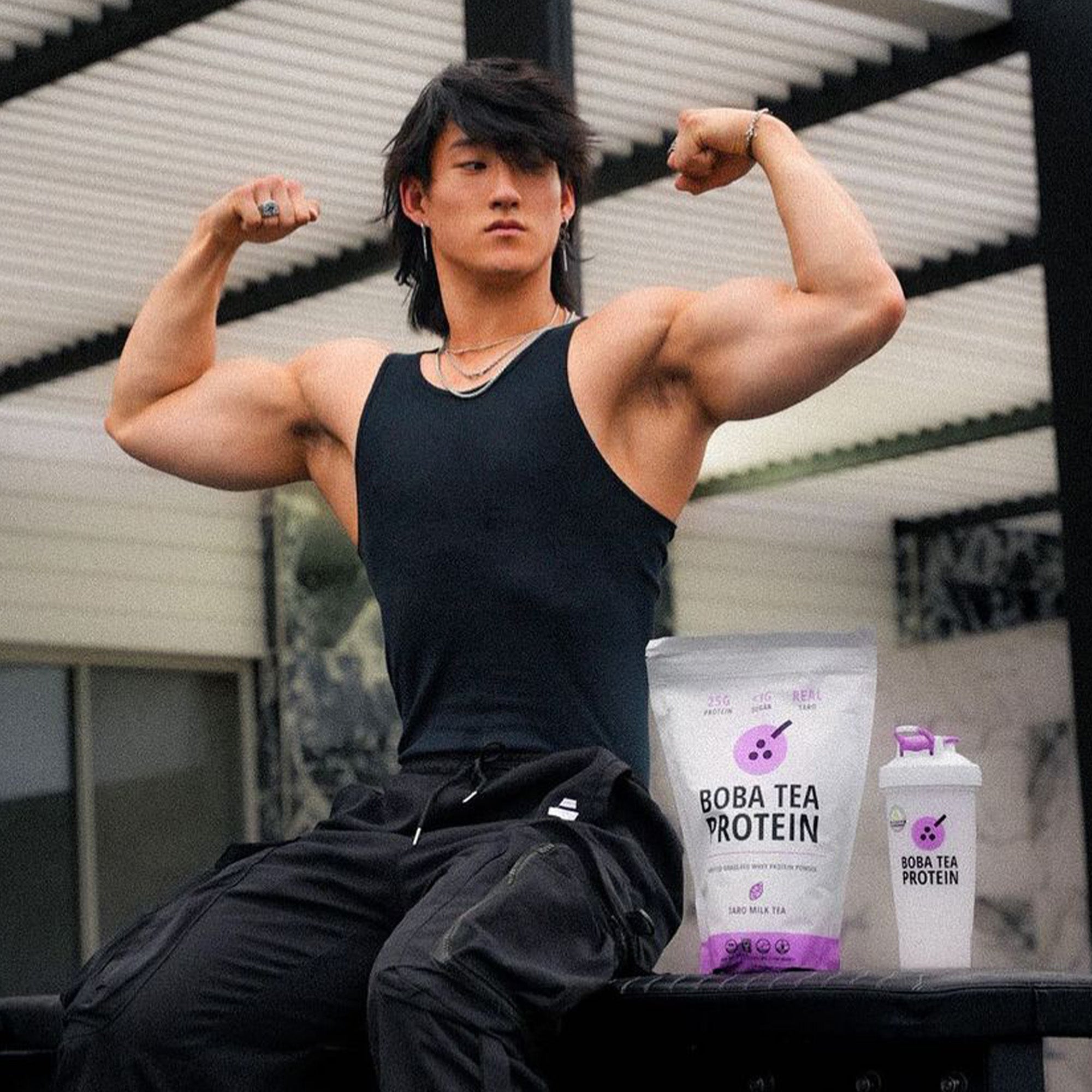 A muscular man in a black tank top poses flexing his arms outdoors, next to a bag of Taro Milk Tea Whey Protein from Boba Tea Protein and a Boba Tea Protein branded shaker bottle in matching Taro purple. 