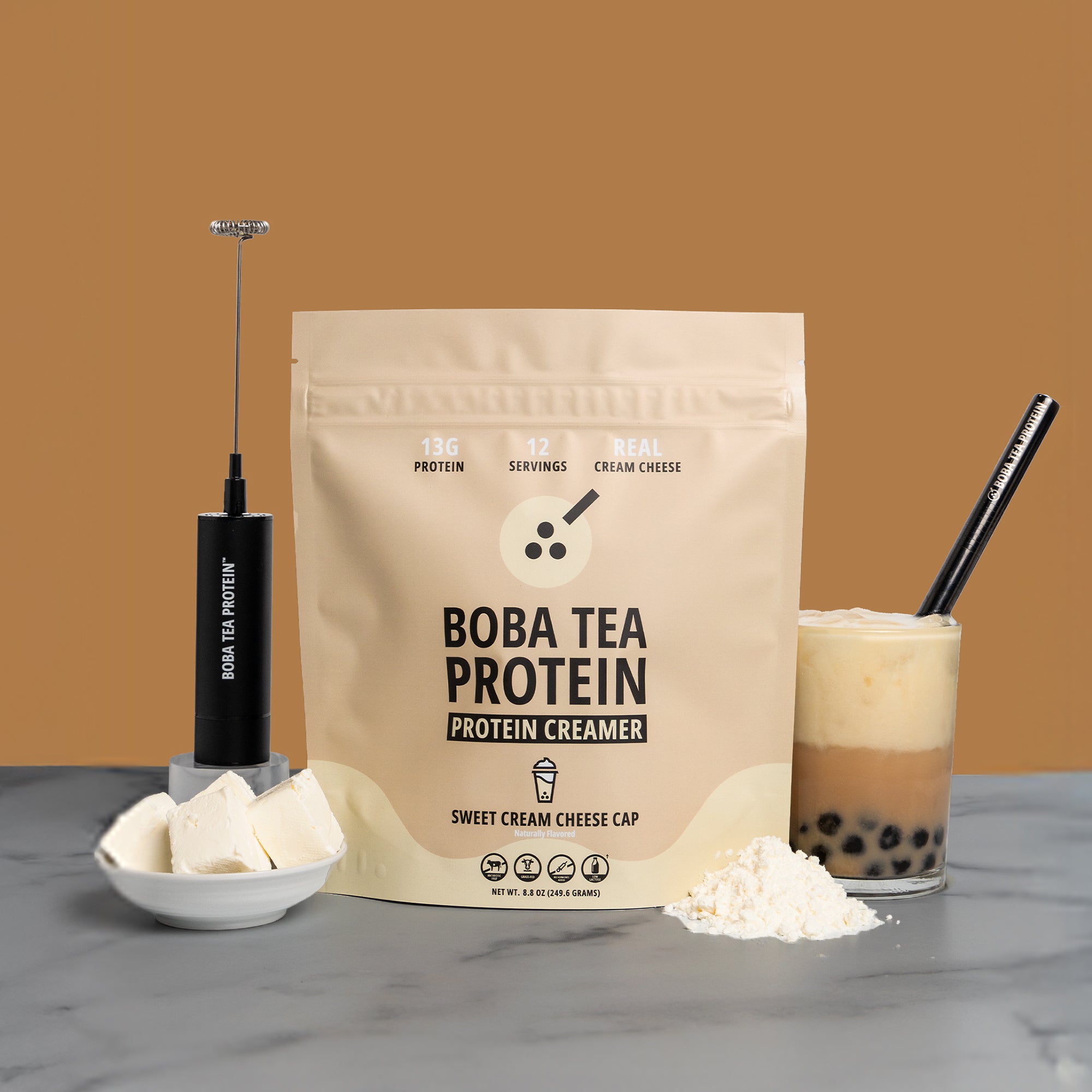 Boba tea protein product with packaging, creamer, and a glass of bubble tea on a brown background.