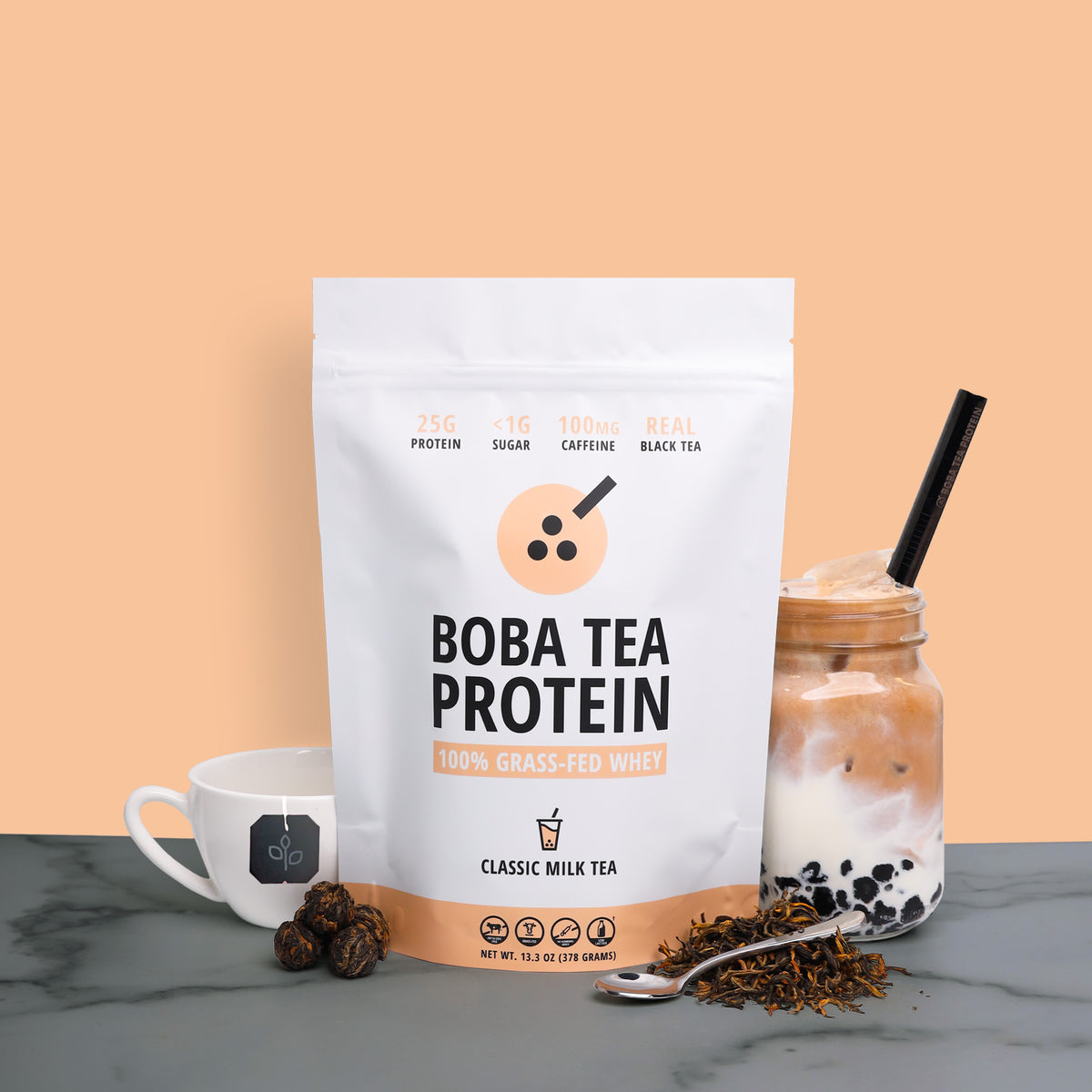 A 10-serving stand-up pouch of Boba Tea Protein powder stands on a gray marble surface against a peach background, styled with a white tea cup, a boba milk tea drink in a glass jar, loose tea leaves, and a spoon.