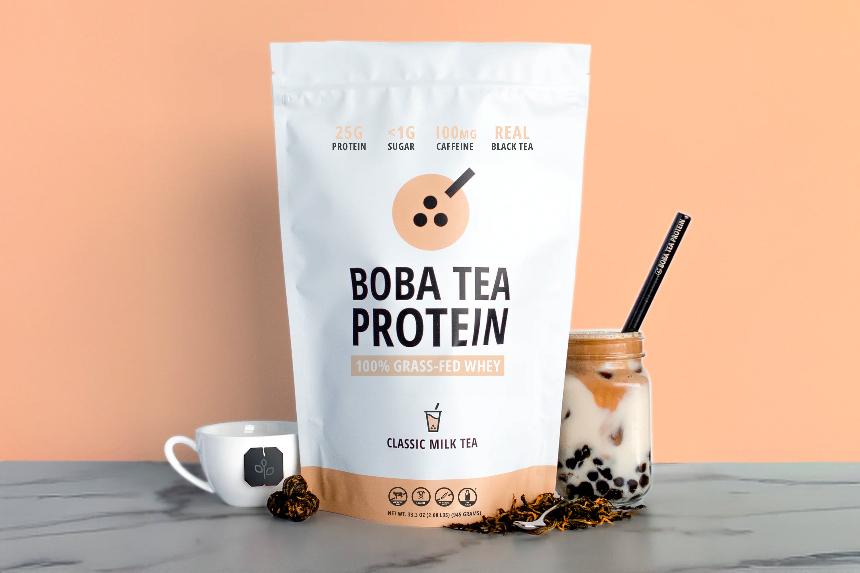 A stand-up pouch of Boba Tea Protein powder stands on a gray marble surface against a peach background, styled with a small white mug, loose tea leaves, and a glass jar of milk tea with a black straw.