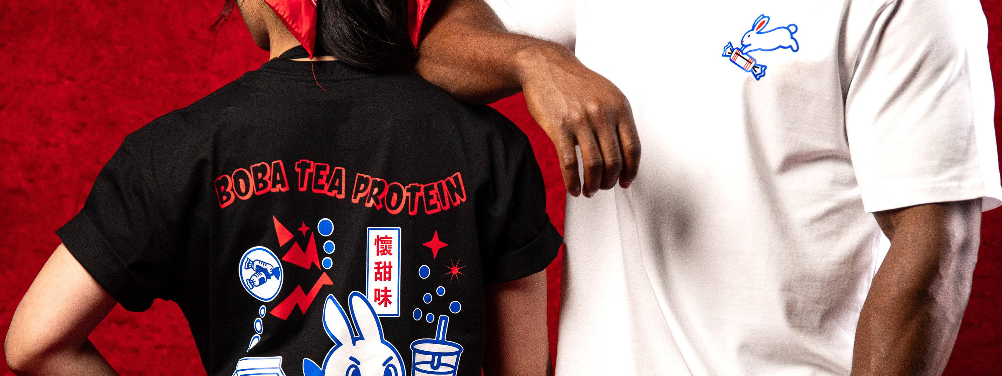 Person wearing a black t-shirt with red text and graphics, standing next to another person in a white t-shirt on a red background.